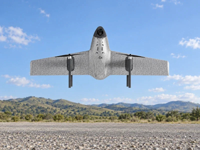 UVH1 in the exact underside reference attitude above an empty gravel survey staging strip with pale green hills beyond, full aircraft visible.