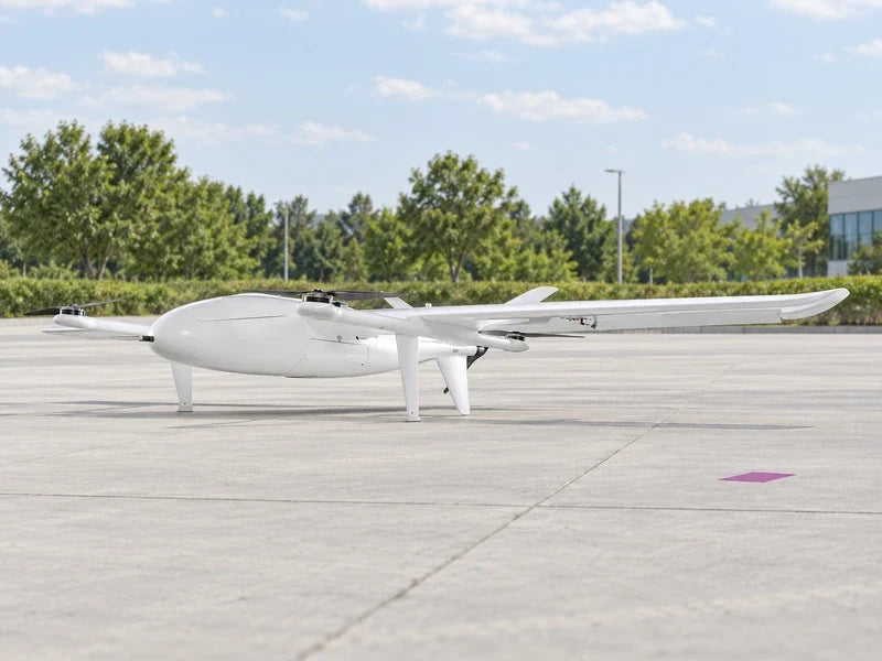 UG25 in exact approved low oblique profile on an empty pale concrete analytics-campus apron with a small magenta floor marker and green trees in distance